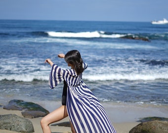 Puede incluir: Una mujer con una bata de playa a rayas azules y blancas con un dobladillo con volantes, de pie en una playa de arena con el océano de fondo.