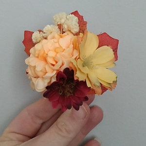 Op de afbeelding: Een klein boeket kunstbloemen met perzik-, gele en bordeauxrode bloemen. De bloemen zijn gerangschikt met gedroogde witte bloemen en bruine bladeren.