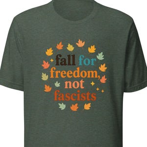 Peut inclure: T-shirt vert foncé avec l'inscription "fall for freedom, not fascists" entourée de feuilles d'automne. Le texte est en orange, bleu et jaune. Le t-shirt est fait d'une matière douce.