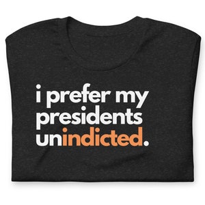 May include: A black t-shirt with the text "i prefer my presidents unindicted." in white and orange.
