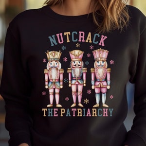 Może przedstawiać: Czarna bluza z humorystycznym wzorem świątecznym przedstawiającym trzy postaci szczupaczy i tekst "Nutcrack the Patriarchy".