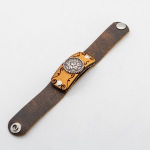 May include: A brown leather bracelet with a decorative silver floral design and a clear rhinestone in the center. The bracelet has a snap closure.