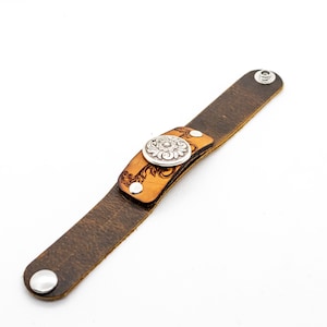 May include: A brown leather bracelet with a silver floral design and a snap closure.