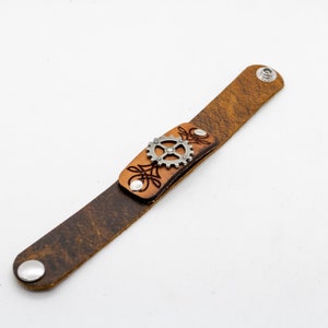 May include: A brown leather bracelet with a silver gear charm. The bracelet has a distressed look and is adjustable.