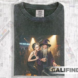 May include: Dark grey Comfort Colors t-shirt with a graphic of a wizard and a woman in a nightclub. The text "THE HOG'S HEAD" is displayed. The tee has a vintage, washed appearance.