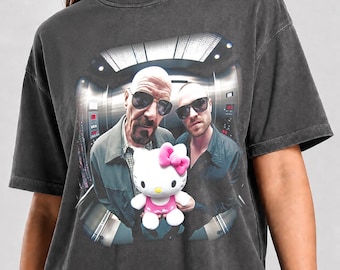 May include: A charcoal gray t-shirt featuring a graphic print of two men in sunglasses standing in an elevator, with a white Hello Kitty plush toy with a pink bow positioned in the foreground. The text Pepper Color is printed at the bottom.