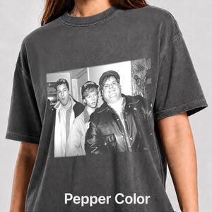 May include: A charcoal grey short-sleeve t-shirt featuring a black and white graphic print of three individuals. The central image shows three people wearing hats and jackets, with the person on the right wearing a leather jacket. The shirt has a vintage-style washed finish.