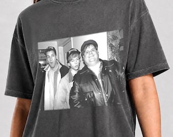 May include: A charcoal gray short-sleeve t-shirt featuring a black and white graphic print of three individuals. The central image shows three people wearing hats and jackets, with the person on the right wearing a leather jacket. The shirt has a vintage-style washed finish.