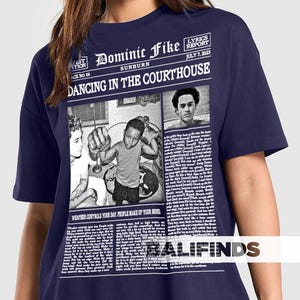 May include: Navy blue t-shirt featuring a newspaper-style graphic with the text "Dominic Fike Sunburn Dancing in the Courthouse." The graphic includes images and the text "Lyrics Report July 7, 2023" and "Weather controls your day. People make up your mind."