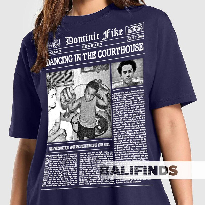May include: Navy blue t-shirt featuring a newspaper-style graphic with the text "Dominic Fike Sunburn Dancing in the Courthouse." The graphic includes images and the text "Lyrics Report July 7, 2023" and "Weather controls your day. People make up your mind."