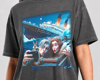 May include: A charcoal gray t-shirt featuring a graphic print of two people taking a selfie in the water with the Titanic ship in the background. The print shows the ship with illuminated smokestacks and icebergs. The text Pepper Color is visible.