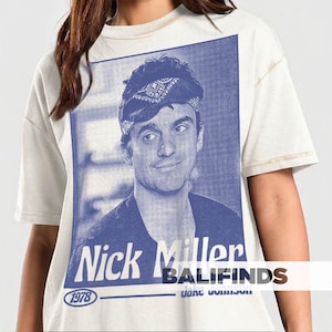 May include: Cream-colored t-shirt featuring a blue and white graphic of Nick Miller. The graphic includes the text "Nick Miller" and the year "1978". The shirt has a vintage, worn look.