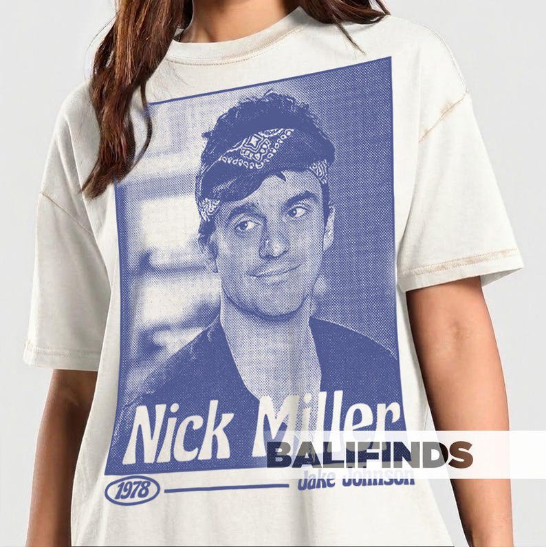 May include: Cream-colored t-shirt featuring a blue and white graphic of Nick Miller. The graphic includes the text "Nick Miller" and the year "1978". The shirt has a vintage, worn look.