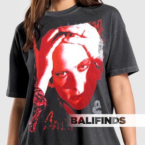 Pu&ograve; includere: T-shirt grigio scuro con grafica rossa e bianca di un volto umano, mani sulla testa. La grafica ha un aspetto invecchiato e vintage. Il testo "BALIFINDS" &egrave; nella parte inferiore della maglietta.