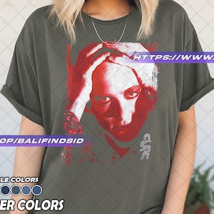 Pu&ograve; includere: T-shirt verde oliva con grafica rossa e bianca di un volto umano con le mani sulla testa. Il testo "HTTPS://WWW.ETSY.COM" &egrave; visibile. Sotto, opzioni di colore e la scritta "PEPPER COLORS".