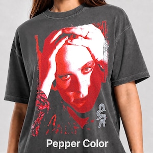 May include: A charcoal gray short-sleeve t-shirt featuring a large, high-contrast red graphic print of a person with hands on their head. The design includes a small white symbol in the lower right corner and the text Pepper Color at the bottom.
