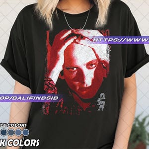 Pu&ograve; includere: T-shirt nero con grafica rossa e bianca di un volto umano, mani sulla testa. La maglietta ha il testo "AVAILABLE COLORS" e "BLACK COLORS" in basso. L'URL del negozio Etsy &egrave; visibile sulla maglietta.