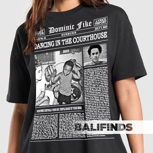 May include: Black t-shirt featuring a newspaper-style graphic with the text "Dominic Fike Sunburn Dancing in the Courthouse." The graphic includes images and the text "Lyrics Report July 7, 2023." The shirt also has the text "Weather controls your day. People make up your mind."