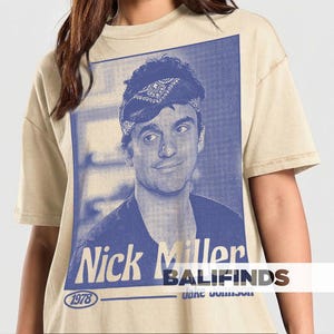 May include: Beige t-shirt featuring a blue and white graphic of a man with a bandana, with the text "Nick Miller" and "1978". The shirt has short sleeves and a crew neck.