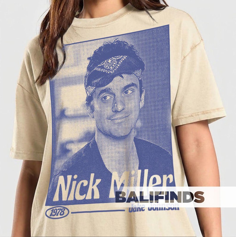 May include: Beige t-shirt featuring a blue and white graphic of a man with a bandana, with the text "Nick Miller" and "1978". The shirt has short sleeves and a crew neck.