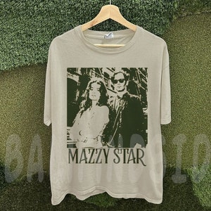 Puede incluir: Una camiseta de color beige claro con una imagen verde y blanca de un hombre y una mujer, con el nombre de la banda "MAZZY STAR" impreso debajo. La imagen es un dise&ntilde;o estilizado y monocrom&aacute;tico. La camiseta est&aacute; colgada en una percha de madera.