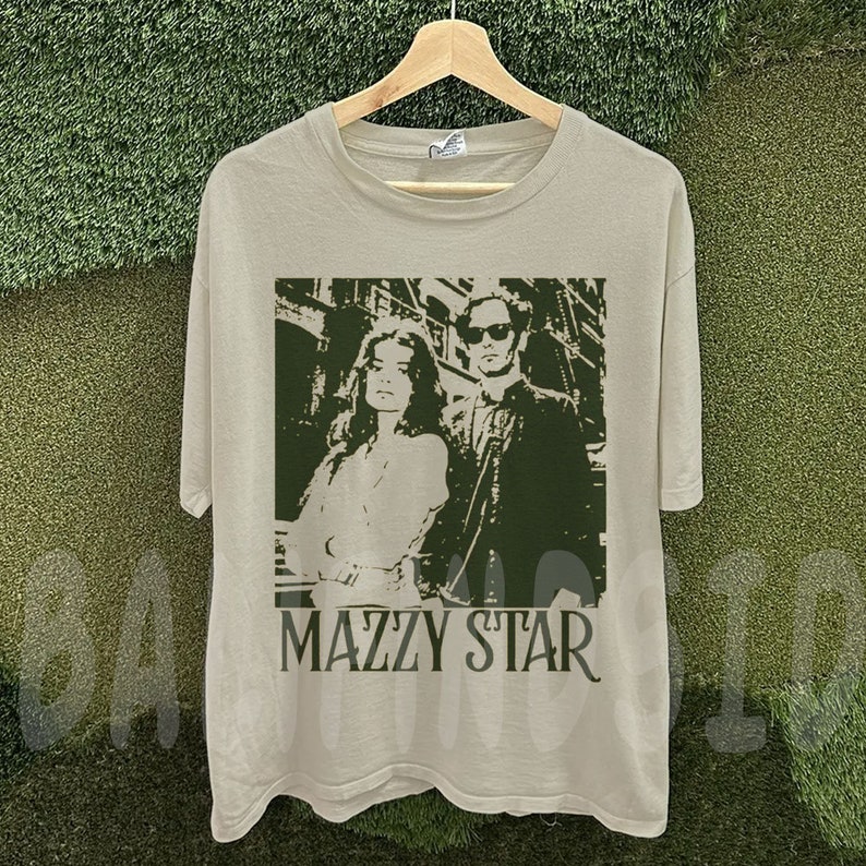 Puede incluir: Una camiseta de color beige claro con una imagen verde y blanca de un hombre y una mujer, con el nombre de la banda "MAZZY STAR" impreso debajo. La imagen es un dise&ntilde;o estilizado y monocrom&aacute;tico. La camiseta est&aacute; colgada en una percha de madera.