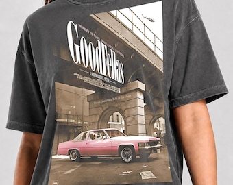 May include: A charcoal gray short-sleeve t-shirt featuring a graphic print of the Goodfellas movie poster. The design displays a pink car parked under a concrete bridge with the text Goodfellas and Three Decades of Life in the Mafia.