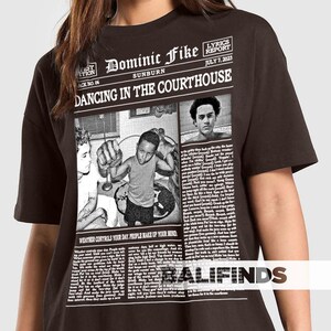 May include: A dark brown t-shirt featuring a newspaper-style graphic with the text "Dominic Fike Sunburn Dancing in the Courthouse." The graphic includes images and text, with the phrase "Weather controls your day. People make up your mind."
