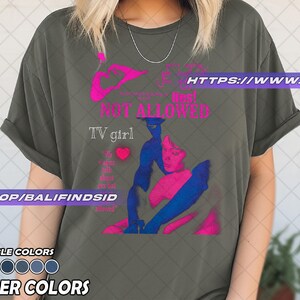 May include: Olive green t-shirt with a graphic design in pink and blue. The design includes the words "NOT ALLOWED" and "TV girl". The shirt also has a heart graphic and the text "I'm alone talk about sex but allowed".