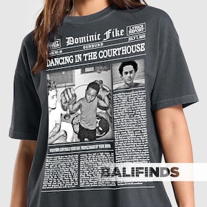 May include: Dark gray t-shirt featuring a newspaper-style graphic with the text "Dominic Fike Sunburn Dancing in the Courthouse." The graphic includes black and white images and text. The shirt also has the text "Weather controls your day. People make up your mind."