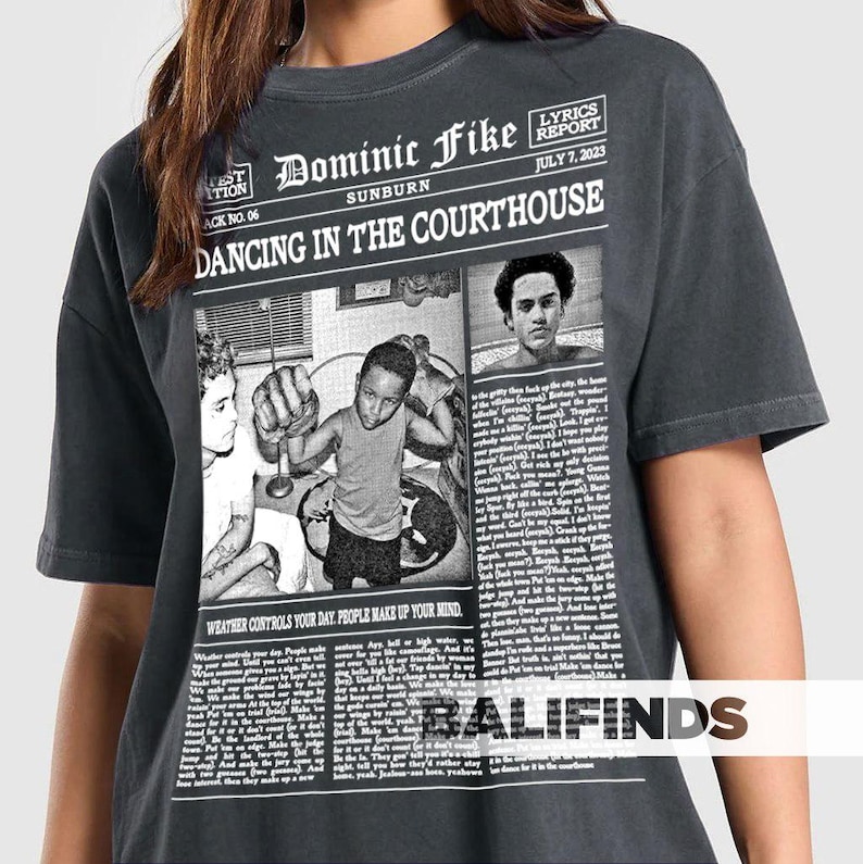 May include: Dark gray t-shirt featuring a newspaper-style graphic with the text "Dominic Fike Sunburn Dancing in the Courthouse." The graphic includes black and white images and text. The shirt also has the text "Weather controls your day. People make up your mind."