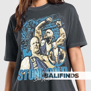 May include: Dark gray t-shirt featuring a graphic of Stone Cold Steve Austin in a wrestling ring. The design includes images of Austin in various poses, with blue and gold accents, and the text "STONE COLD" and "Steve Austin".