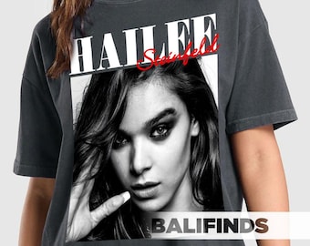 May include: Dark gray t-shirt featuring a black and white image of Hailee Steinfeld. The shirt has the name "HAILEE" in white block letters and "Steinfeld" in red script. The word "BALIFINDS" is printed in white at the bottom of the image.