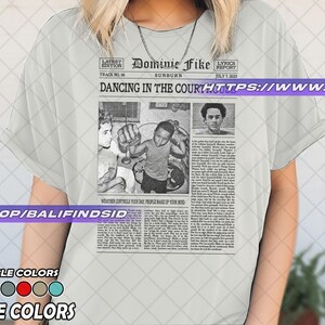 May include: White t-shirt featuring a newspaper-style graphic with the headline "Dancing in the Court" and the name "Dominie Fike." The graphic includes black and white images and text. Available in various colors.