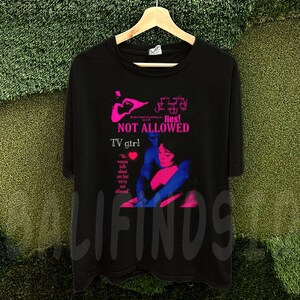 May include: Black t-shirt with a graphic design featuring the text "NOT ALLOWED" and "TV girl" in pink and white. The design includes an illustration of two people embracing. The shirt is hanging on a wooden hanger.