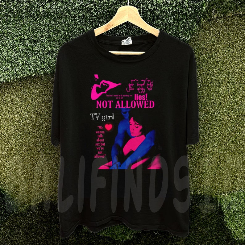 May include: Black t-shirt with a graphic design featuring the text "NOT ALLOWED" and "TV girl" in pink and white. The design includes an illustration of two people embracing. The shirt is hanging on a wooden hanger.