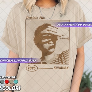 May include: Ivory-colored t-shirt featuring a sepia-toned graphic of a person shielding their eyes, with the text "Dominic Fike" above and "Sunburn 2023" below. The shirt also displays "Available Colors" and "Ivory Colors".
