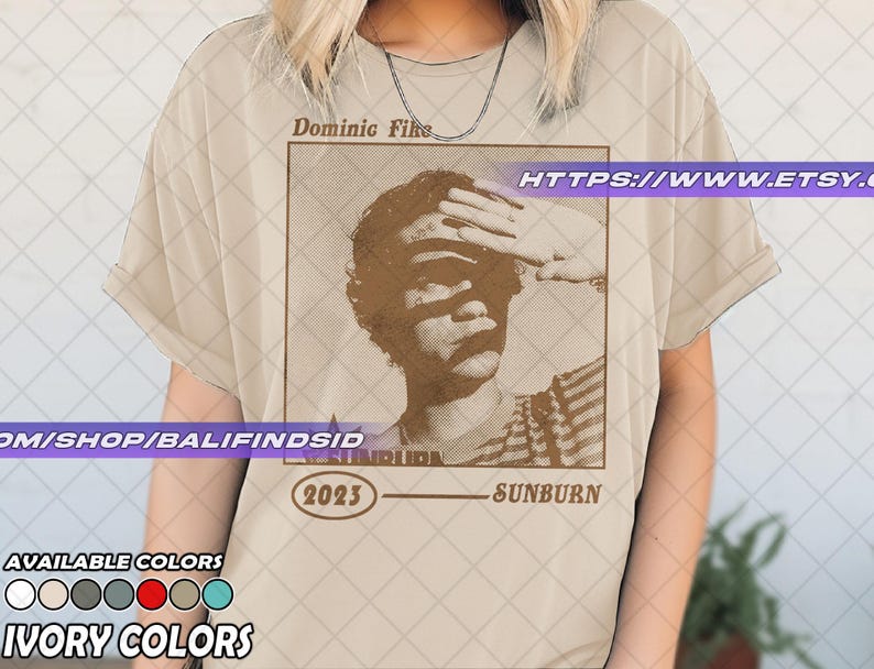 May include: Ivory-colored t-shirt featuring a sepia-toned graphic of a person shielding their eyes, with the text "Dominic Fike" above and "Sunburn 2023" below. The shirt also displays "Available Colors" and "Ivory Colors".