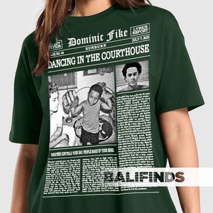 May include: Dark green t-shirt featuring a newspaper-style graphic with the text "Dominic Fike Sunburn Dancing in the Courthouse." The graphic includes images and the text "Weather controls your day. People make up your mind."