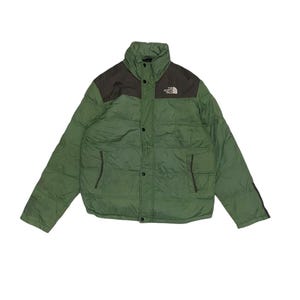 May include: A green puffer jacket with a high collar and a dark brown panel across the upper chest and shoulders. The jacket features a front snap button closure, two side pockets, and the white The North Face logo on the upper left chest.