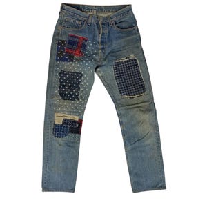 May include: Light blue denim jeans featuring a button fly and multiple decorative fabric patches. The patches include various patterns such as red and blue plaid, dark blue geometric prints, and striped fabric, all stitched onto the legs.