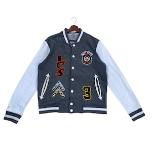May include: A dark gray varsity jacket with white sleeves, featuring white snap buttons, striped ribbed cuffs, and a collar. Patches include the text LCS, a rooster logo with the text WIT COQ LE COQ SPORTIF, the number 3, and Le Coq Sportif branding.