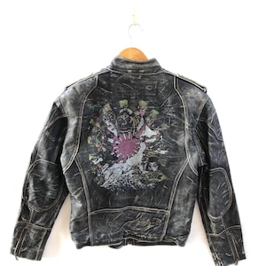 May include: A black leather jacket with a distressed, washed-out finish. The back features a large, colourful graphic print depicting a stylised warrior figure, a rising sun, and wave patterns. The jacket includes a stand-up collar and zippered cuffs.