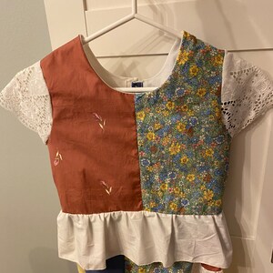 May include: A patchwork dress with a brown and floral print top, white lace sleeves, and a white ruffle trim. The dress has a blue and brown patchwork skirt.