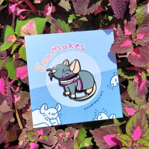 Peut inclure: Une épingle en émail noir et gris représentant une souris de dessin animé portant une écharpe rayée violette, blanche et noire. L'épingle est sur un fond bleu avec le texte "EonMakes" et "eondraws.art" en blanc.