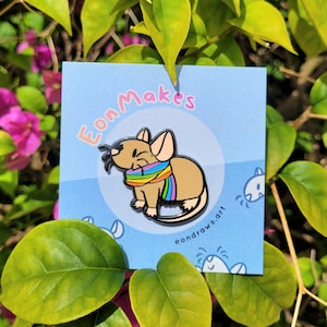 May include: A colourful enamel pin featuring a cartoon mouse wearing a rainbow scarf. The pin is on a white card with the text "Eon Makes" and "eondraws.art" in black.