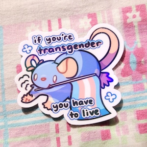 Puede incluir: Una pegatina con un ratón azul de dibujos animados sosteniendo una bandera del orgullo transgénero. La pegatina dice "si eres transgénero, tienes que vivir".