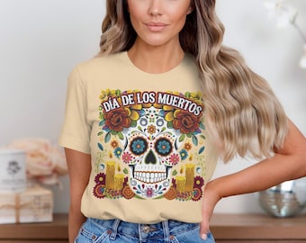 May include: A light beige t-shirt featuring a colorful sugar skull design with the text "Día de los Muertos". The skull is decorated with flowers and candles, celebrating the Day of the Dead.