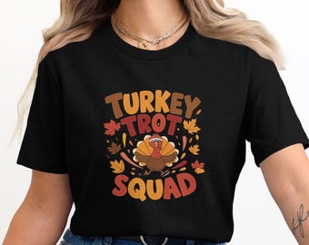 May include: Black t-shirt with a colorful graphic that reads "TURKEY TROT SQUAD." The design features a cartoon turkey, autumn leaves, and text in shades of brown, orange, and red. A festive apparel item.