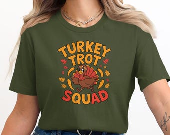 May include: Olive green t-shirt with a colorful graphic. The graphic features a cartoon turkey wearing a headband and running, with the words "TURKEY TROT SQUAD" in orange and red. Autumn leaves surround the turkey.